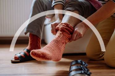 Person helps another put on an orange sock in a room with wooden flooring.