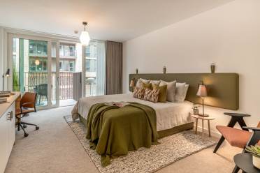 Modern bedroom with a large bed, green headboard, and neatly arranged pillows. There is a side table with lamps, a desk on the left, and a balcony accessible through sliding glass doors in the background.