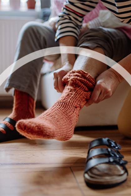Person helps another put on an orange sock in a room with wooden flooring.