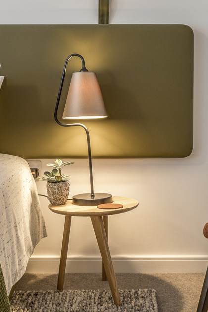 A modern bedroom features a neatly made bed, a bedside table with a lamp and a small plant, and a brown upholstered chair. The room has a minimalist design with earthy tones.