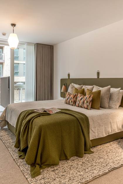 Modern bedroom with a large bed, green headboard, and neatly arranged pillows. There is a side table with lamps, a desk on the left, and a balcony accessible through sliding glass doors in the background.