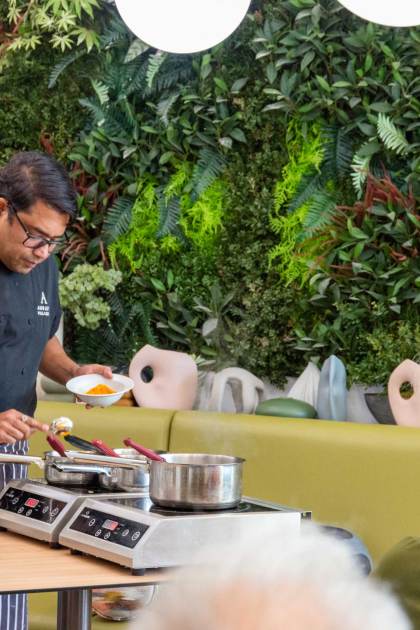 A chef demonstrates cooking at a table with ingredients, pans, and a hot plate, while people watch. The background features a lush, green wall.