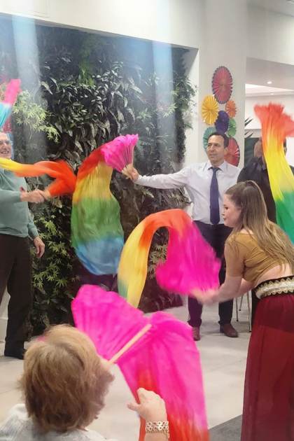 A group of people wave colorful rainbow scarves while others watch in a decorated indoor space with tables and greenery.
