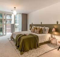 A modern bedroom with a large bed, green and white decor, a desk, chairs, and a window opening to a balcony.