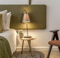 A modern bedroom features a neatly made bed, a bedside table with a lamp and a small plant, and a brown upholstered chair. The room has a minimalist design with earthy tones.