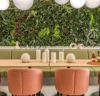 Dining area with a wooden table, pink chairs, and green cushioned bench. Decorative vases on the table. Green vertical garden wall and round ceiling lights in the background.