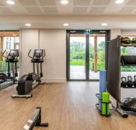 A gym room with exercise equipment, including treadmills, exercise bikes, kettlebells, medicine balls, and stability balls. A glass door provides a view of an outdoor area.