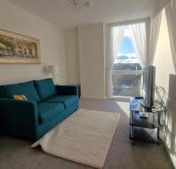 A bright living room with a teal sofa, a white rug, two side tables with lamps, a wall painting, and a TV on a stand. Sunlight streams in through a large window with sheer curtains.