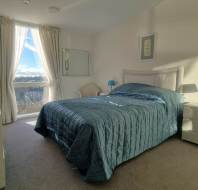 A tidy bedroom with a blue quilted bedspread, white nightstands with blue lamps, and a window with sunlight streaming in through sheer curtains.