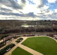 A grassy courtyard with pathways and benches is surrounded by trees and leads to a river. Residential buildings are visible on the right, and the sky is cloudy.