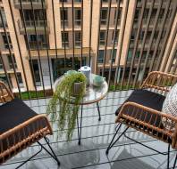Two rattan chairs with cushions face a glass table on a balcony. The table holds a plant, cups, and a teapot. The view includes a brick building with multiple windows.