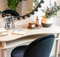 Vanity table with mirror, decorative plant, candle, framed photo, book, and bottle on tray. Black chair in front.