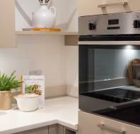 Modern kitchen corner with a built-in oven, light countertops, a cookbook, potted plant, and ceramic containers.