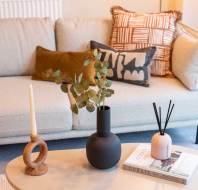 A beige sofa with decorative cushions, and a coffee table with a candle, plant in a black vase, and a reed diffuser atop two books.
