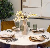 A round dining table set for four with rustic plates, green glasses, and napkins. A vase with white flowers is in the center. Greenery and abstract art decorate the background.