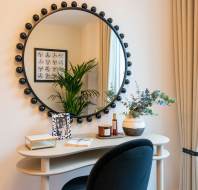A stylish home office with a curved desk, black chair, and a round mirror reflecting a plant and wall art. A vase, candles, and books are on the desk. Beige curtains frame a window.