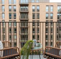 Two wicker chairs around a small table with a kettle and cups on a balcony, facing a modern apartment building.