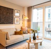 Modern living room with a beige sofa, patterned cushions, and a wooden coffee table. Art prints hang on the wall. A lamp illuminates the space. Balcony with seating is visible through glass doors.
