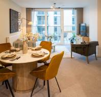 Modern open-plan living room and dining area with a round table, mustard chairs, and a TV. Large windows in the background offer a view of a balcony and buildings.
