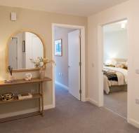 A hallway with a wooden console table, round mirror, and lamp. Two open doors lead to a bedroom and another room. Beige walls and carpeted floor.