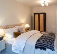Modern bedroom with a neatly made double bed, patterned headboard, bedside tables with lamps, a dark wardrobe, and a ceiling light. A folded throw is placed at the foot of the bed.
