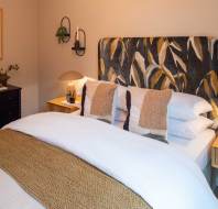 A cozy bedroom with a patterned headboard, neatly made bed, two side tables with lamps, and a dresser with decor. Walls are beige, featuring framed art and a wall-mounted plant.