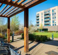 Wooden pergola with wicker chairs overlooks a landscaped garden in front of a modern multi-story apartment building. Clear blue sky in the background.