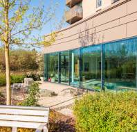 A modern building with large glass windows is adjacent to a landscaped area featuring a tree and shrubs, with a wooden bench in the foreground.