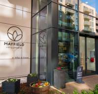 Entrance of Mayfield Villages residential building, featuring glass doors, signage, and planters with flowers.