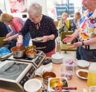People serving themselves food from a buffet table with various dishes, bowls, and cooking equipment in a casual setting.