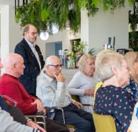 A group of elderly people sitting and listening in a room decorated with plants.