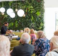 A chef demonstrates cooking techniques to an audience seated in a modern room with a green vertical garden and spherical lights.