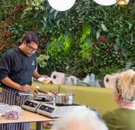 A chef demonstrates cooking at a table with ingredients, pans, and a hot plate, while people watch. The background features a lush, green wall.
