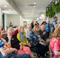 A group of elderly people seated indoors, with some staff members in the background near a reception area. Green plants hang from the ceiling above.