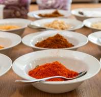 White bowls filled with various colorful spices and herbs are arranged on a wooden surface, each with a metal spoon.