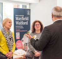 People standing indoors having a conversation near a banner for Mayfield Watford Retirement Living.