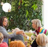 A woman in a gray blazer hands a glass to an older woman wearing a colorful sweater. They are in a room with a green plant wall and other seated people.
