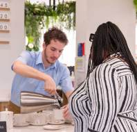 A man in a blue shirt pours coffee from a pot into a cup for a woman in a striped shirt at a café counter.