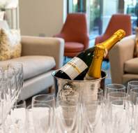 Champagne bottles in an ice bucket surrounded by empty champagne flutes on a table in a lounge area with sofas and chairs in the background.