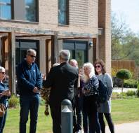 A group of people stand outside a brick building, engaged in conversation, with a grassy area and trees in the background.