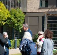 A group of people are engaged in conversation outdoors near a building, with trees and a trellis in the background. Several are wearing jackets, and one person gestures as they talk.