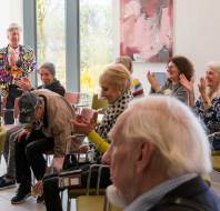 A group of people sitting in a room with chairs, some clapping and smiling. A person in a colorful jacket stands at the back. There are large windows and a red abstract painting on the wall.