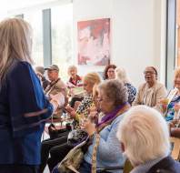 A group of older adults are seated and smiling in a bright room while a woman with long gray hair stands speaking to them.