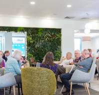 A group of people sitting in a lounge area with green chairs and a plant wall, engaging in conversation.