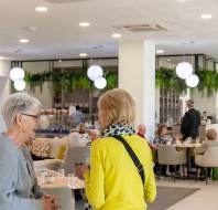 Two women are talking in a modern café with spherical lights, greenery, and people seated at tables in the background.