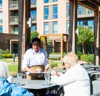 A server in a white shirt and beige apron attends to two seated elderly customers at an outdoor restaurant, with apartment buildings and greenery in the background.