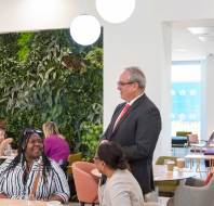 A man in a suit smiles while talking to two seated women in a brightly lit room with a green plant wall.