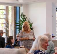 People sitting and chatting in a bright, modern lounge with large windows and potted plants. A woman stands holding a cup, while others are seated on sofas.