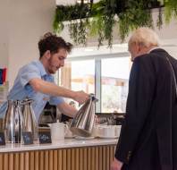 A barista pours a beverage for a customer at a cafe counter with metal jugs labeled for milk, tea, and coffee. Greenery hangs above.
