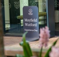 Sign reading "Mayfield Watford Experience Day" in front of a modern building entrance, with flowers in the foreground.